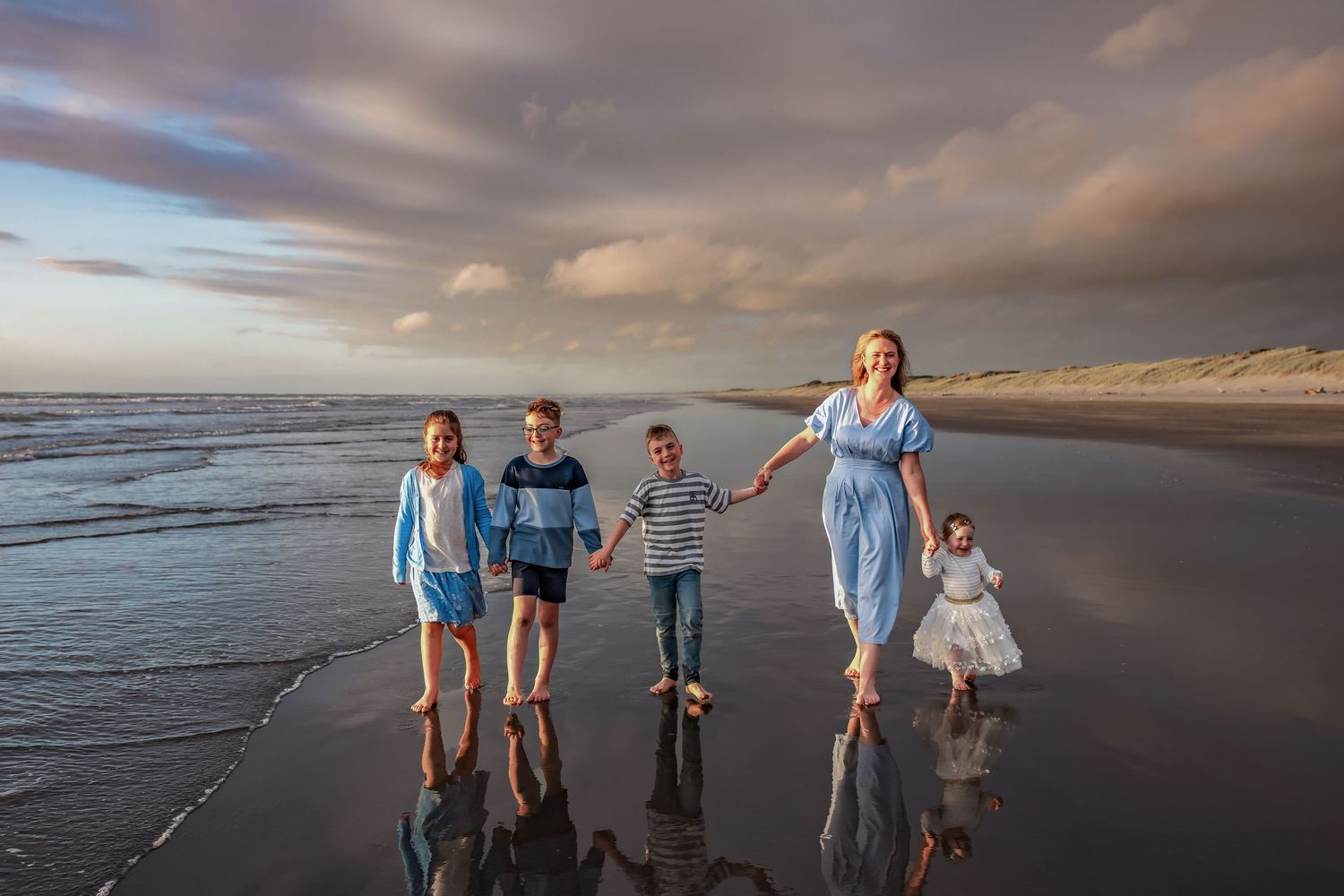 Family enjoying a sunset photoshoot on a cloudy beach with dramatic skies and ocean waves in the background.