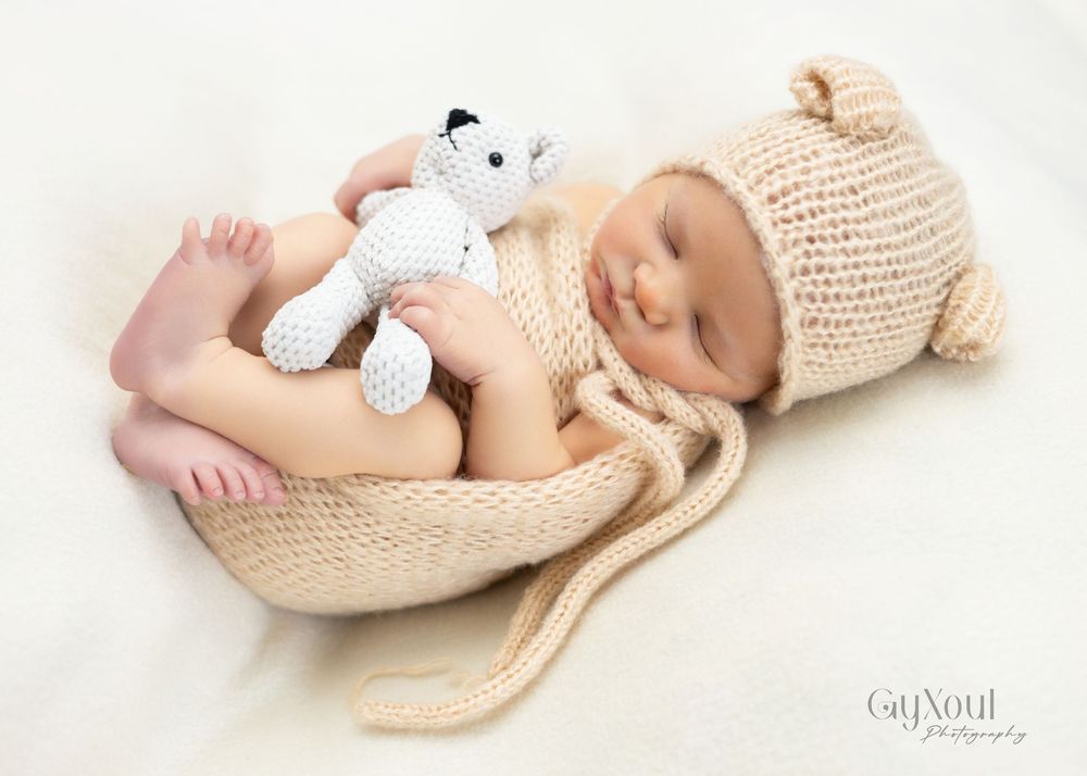 Sleeping baby wearing knitted beige hat lying on soft white blanket with stuffed toy companion.
