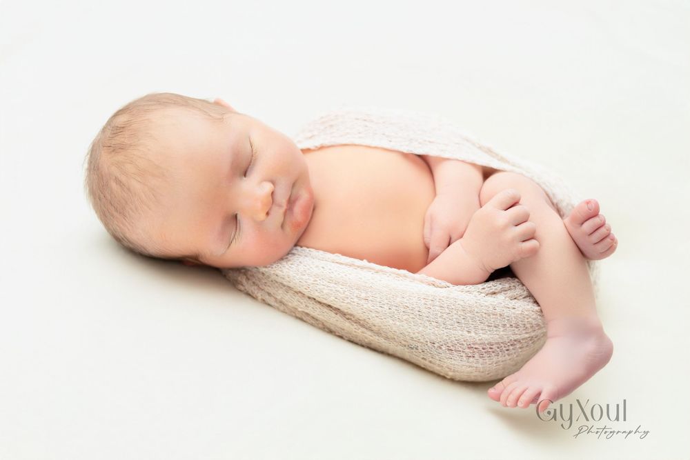 A sleeping newborn baby lies peacefully in a soft beige wrap on a white background.