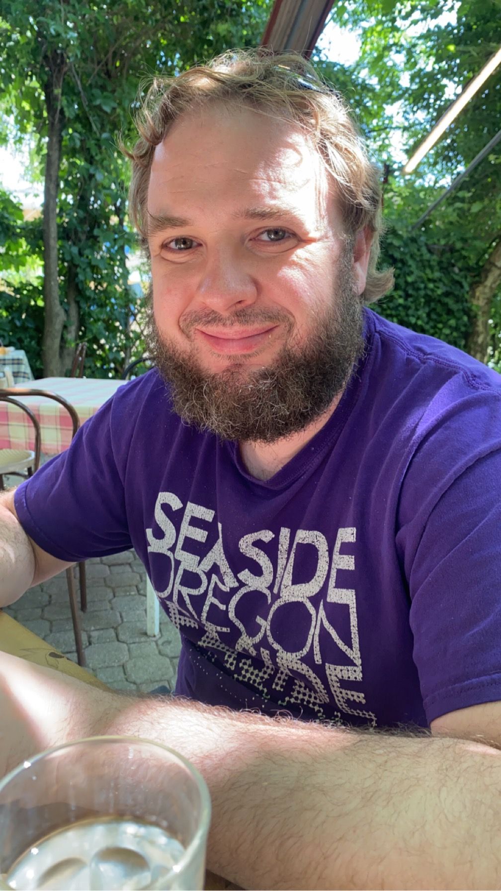 A series of selfies showing someone wearing a purple t-shirt with text while sitting at a table outdoors.