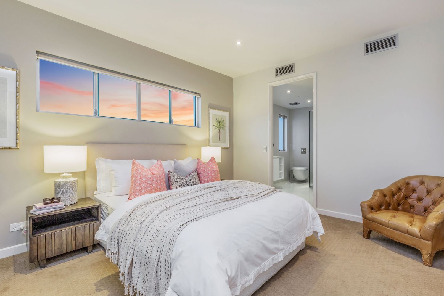 Bright bedroom with queen bed, pink cushions, leather armchair, and ensuite bathroom access.