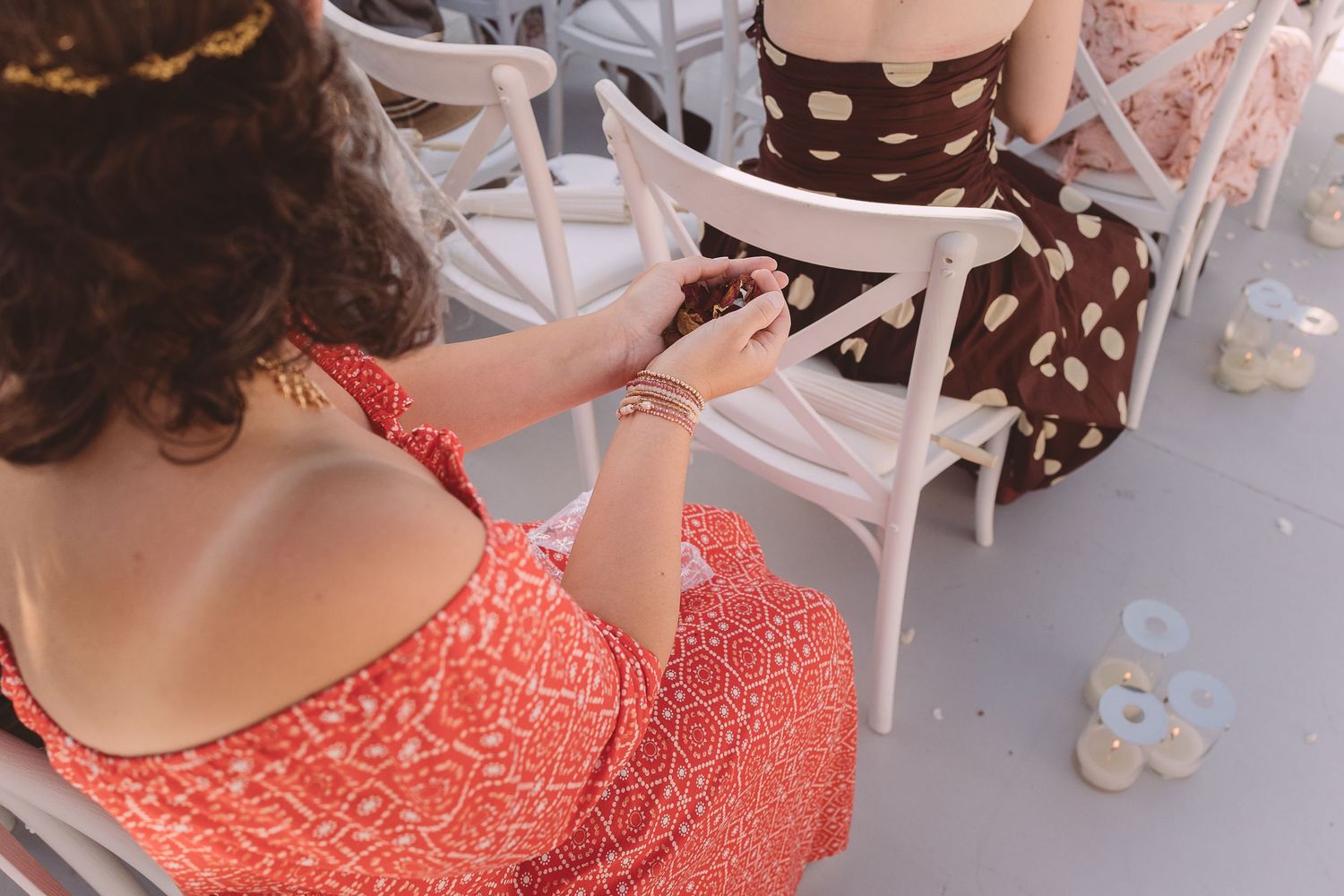 A wedding guests wearing a coral lace dress holding confetti in her hand sitting on a white wooden chair.
