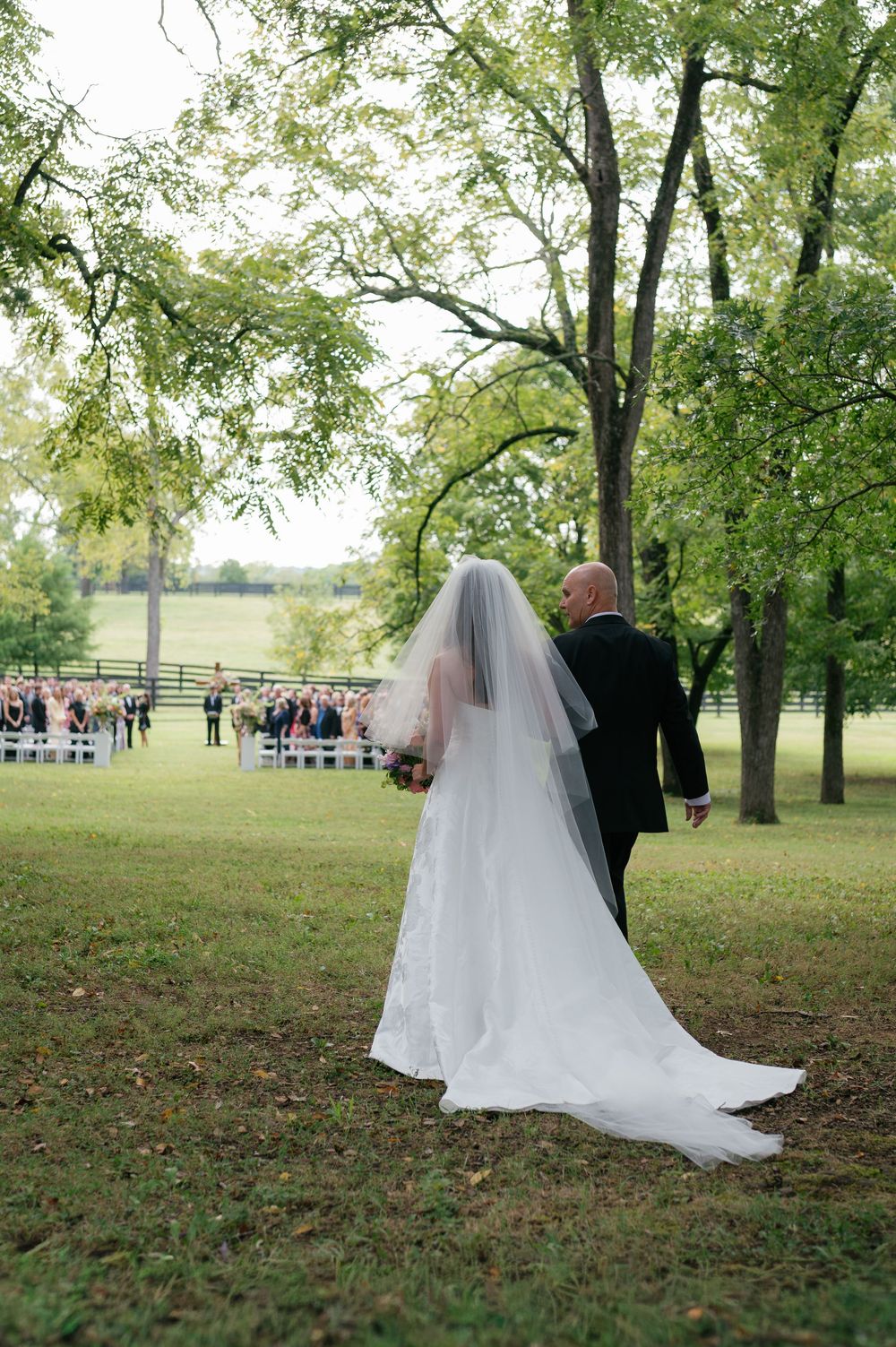 A wedding ceremony takes place outdoors under tall trees with guests seated on the lawn.
