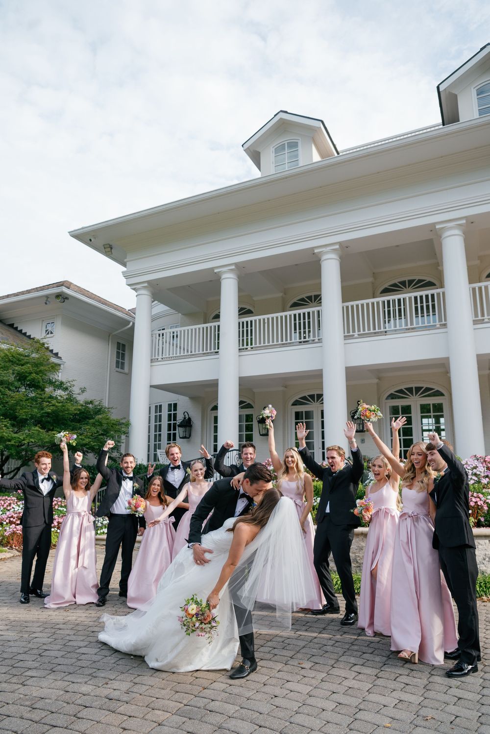 Wedding party celebrates with raised arms in front of elegant white columned mansion.