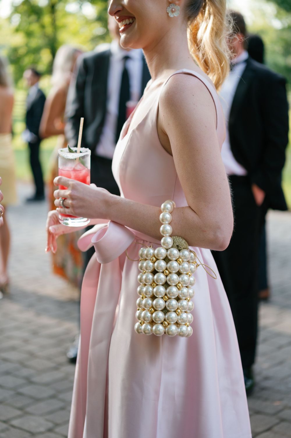Wedding guest in pink dress with pearl accessories holding a cocktail at outdoor reception.