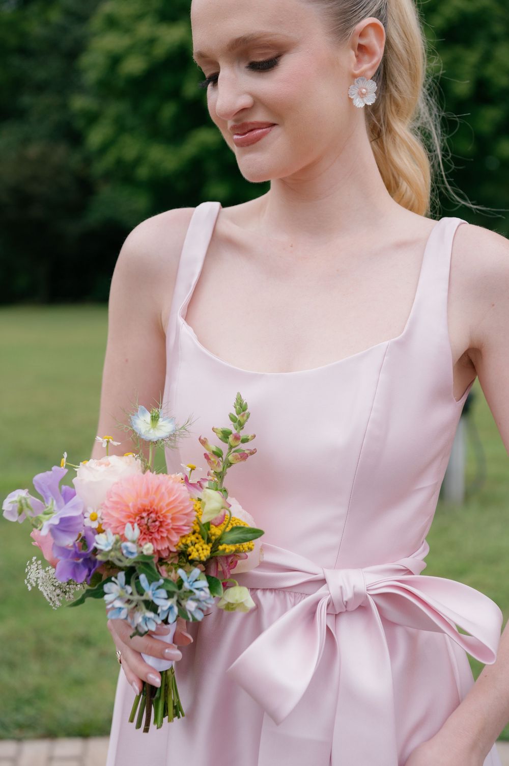 A person in a soft pink dress with a bow detail holds a colorful bouquet of flowers against a green outdoor background.