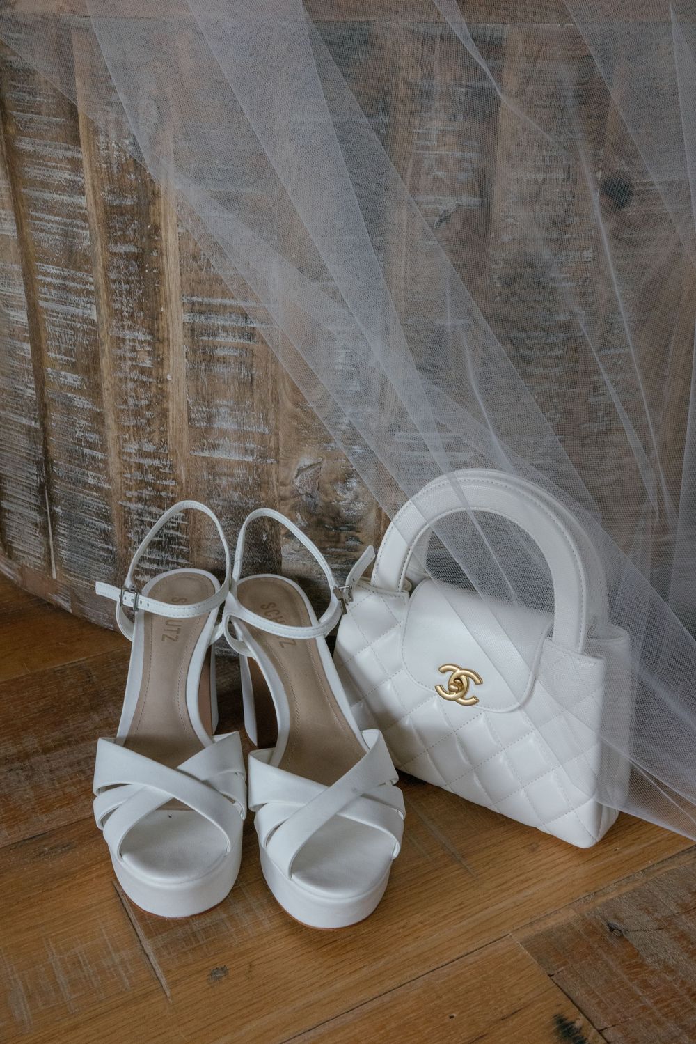 Elegant white bridal shoes and a small white Chanel purse arranged on a wooden floor with delicate tulle fabric.