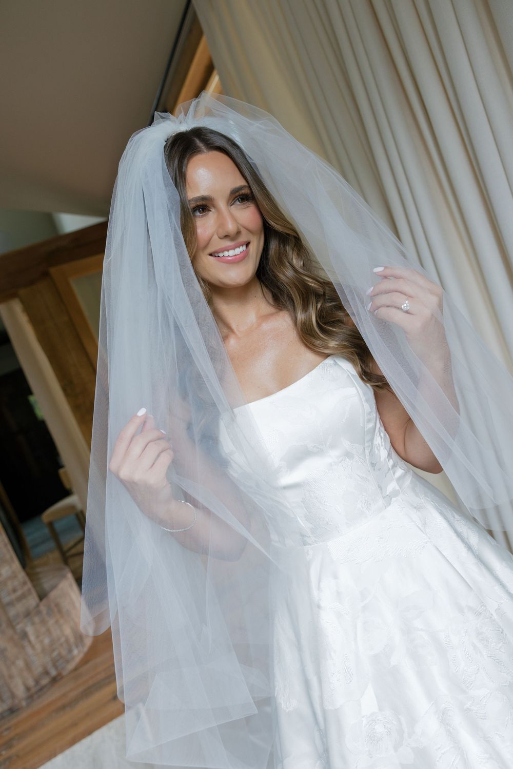 A bride in a white wedding dress and veil poses behind sheer curtains in an indoor setting.