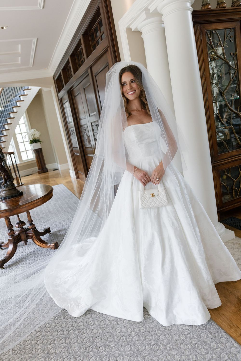 A bride in an elegant white wedding gown with a flowing veil poses in a classic indoor setting.