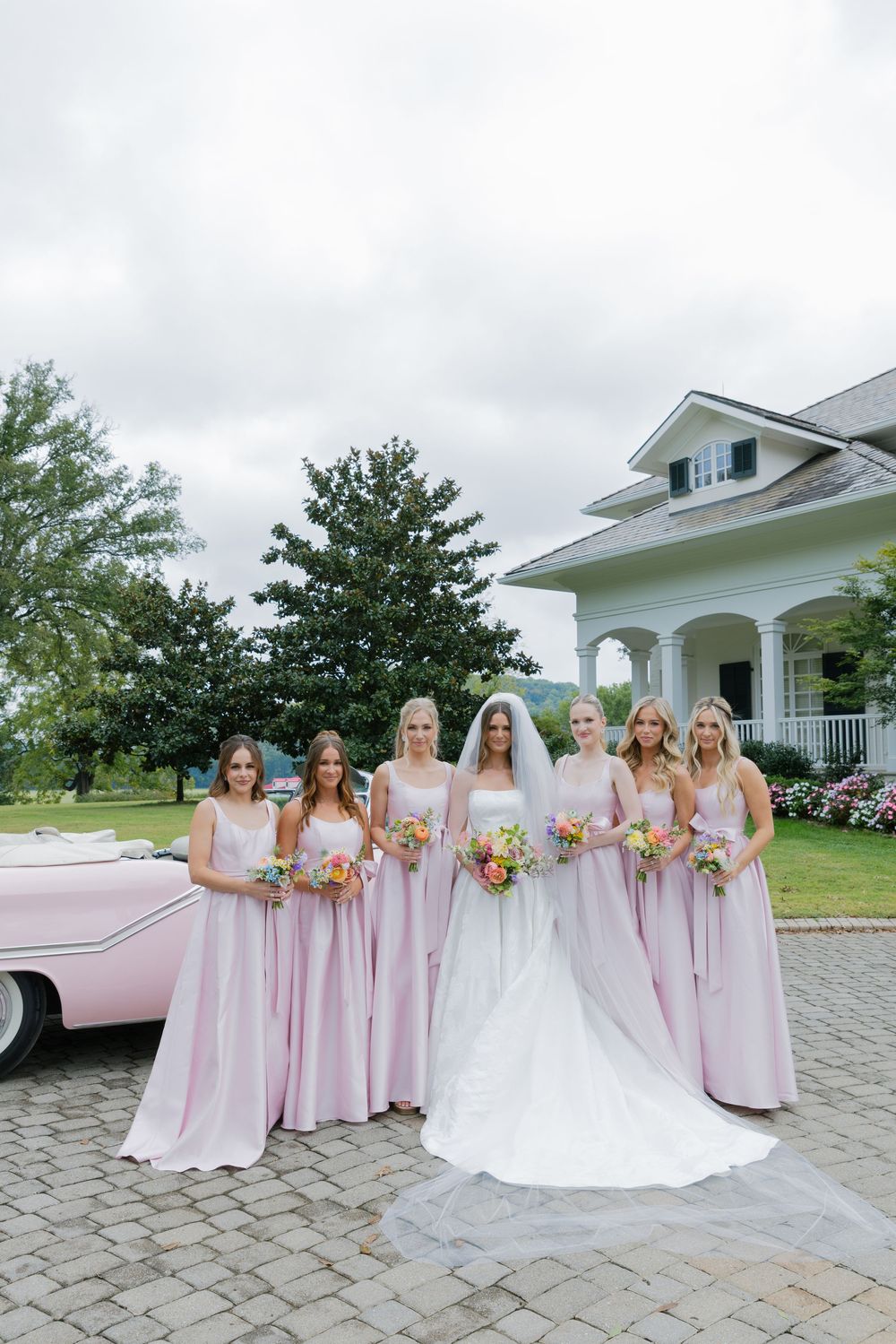 A bridal party in pink dresses poses with the bride in white outside a classic white mansion.