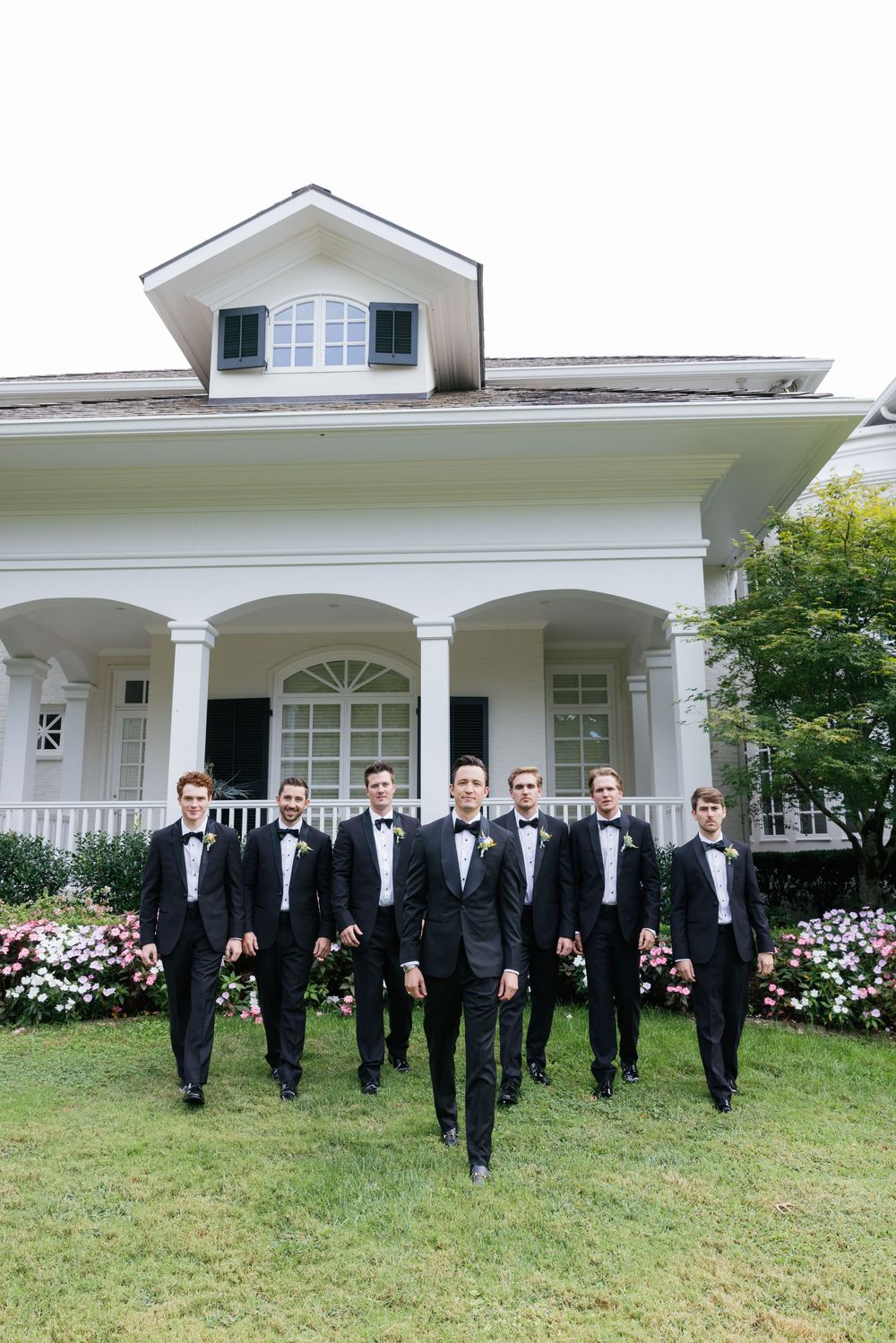 Groomsmen in black tuxedos walk in formation in front of a white colonial house with flowering gardens.