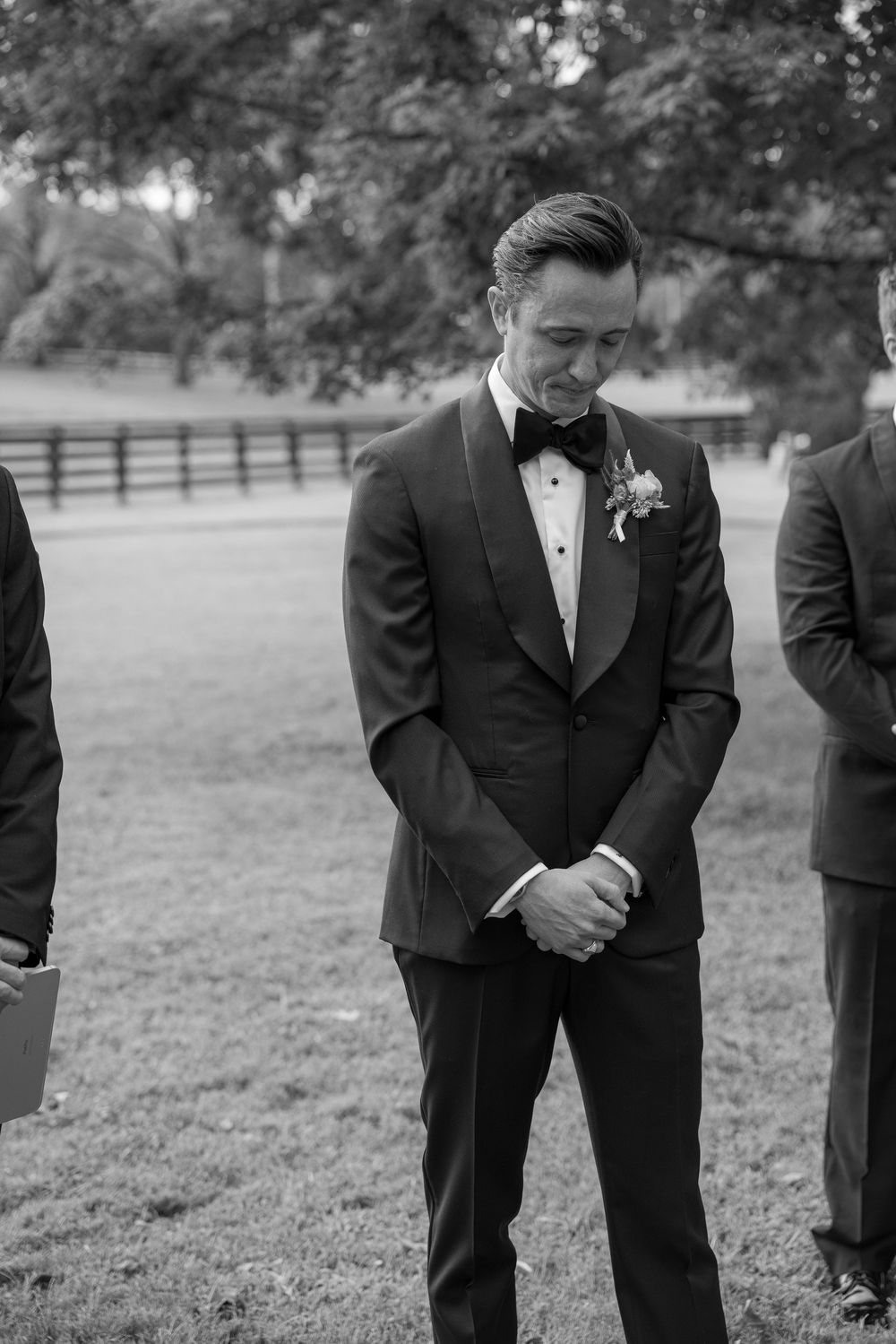 Groomsmen in black suits stand in formation on grassy field in black and white photography.