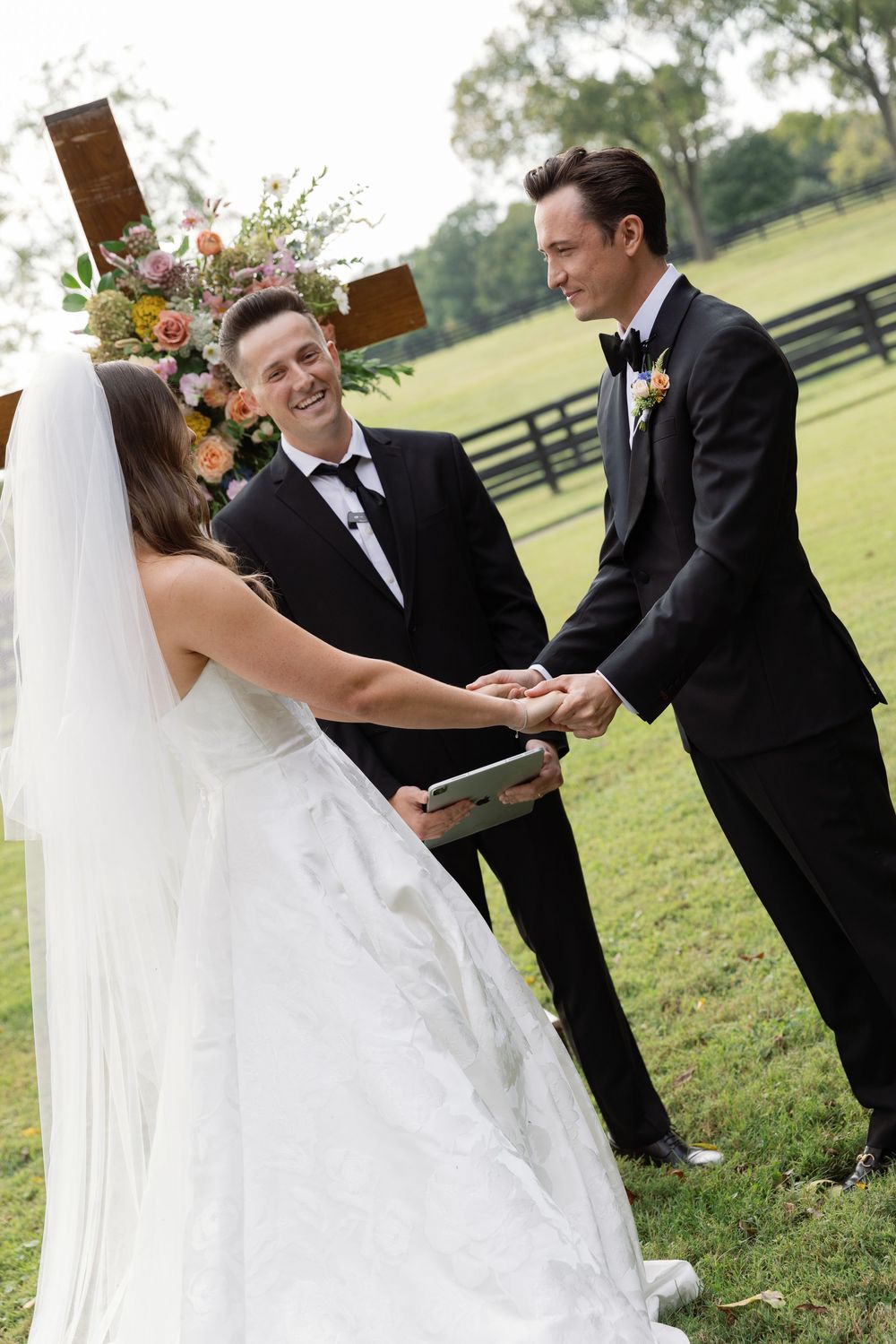 A wedding ceremony takes place outdoors with the bridal party in black suits on a grassy field.