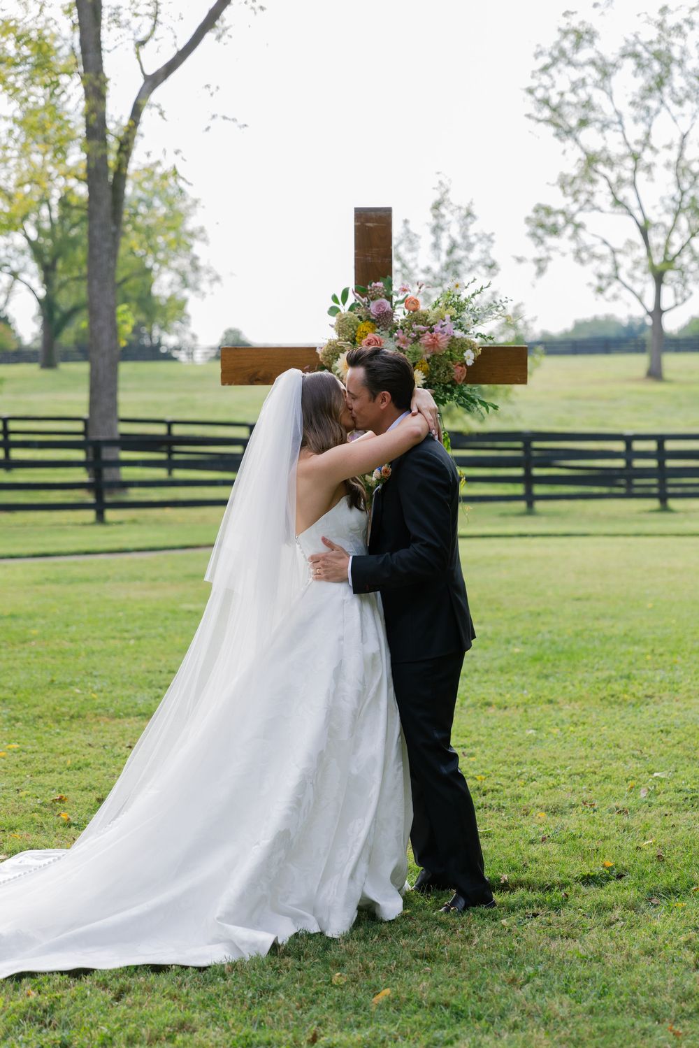 A romantic wedding couple shares a kiss in a scenic outdoor field with wooden fences in the background.