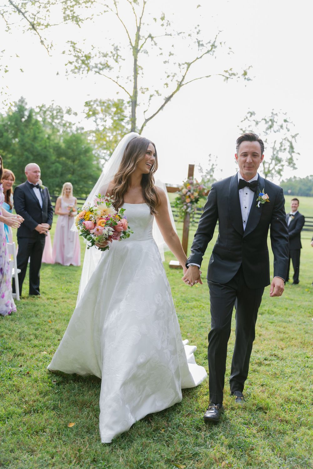 The wedding party walks across a grassy field during an outdoor celebration.