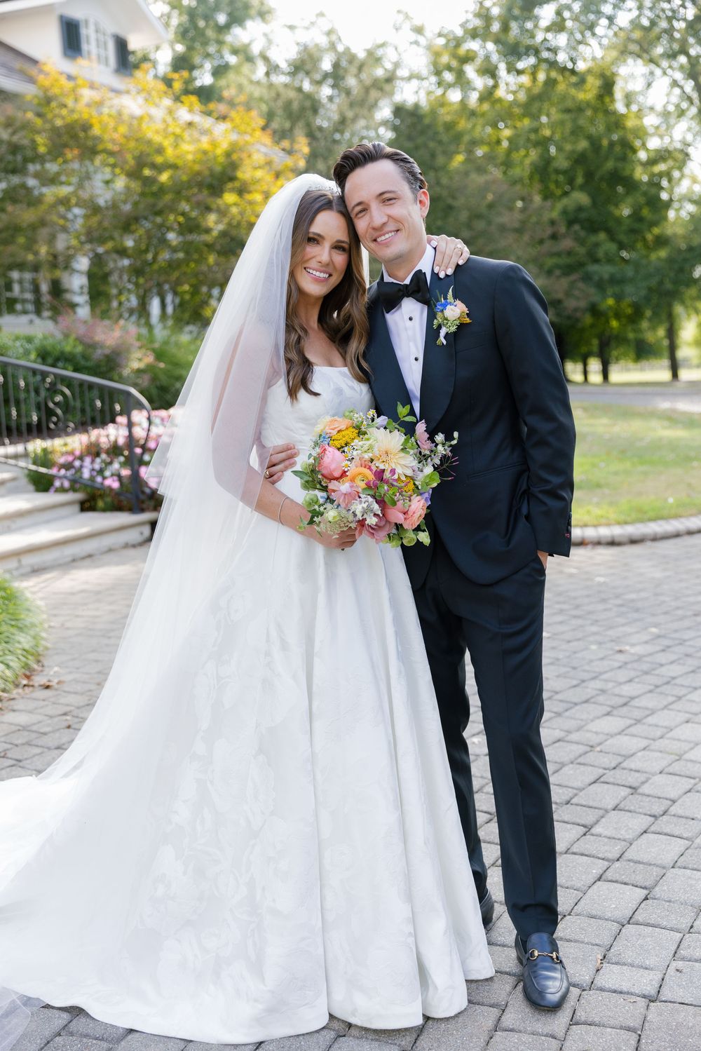 A bride in a white gown and groom in a tuxedo pose together on a stone path with yellow flowers in the background.