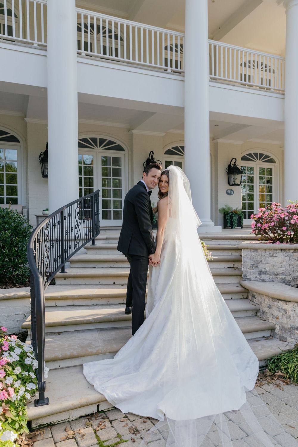 Wedding couple poses on elegant white columned porch with stone steps and flowering gardens.