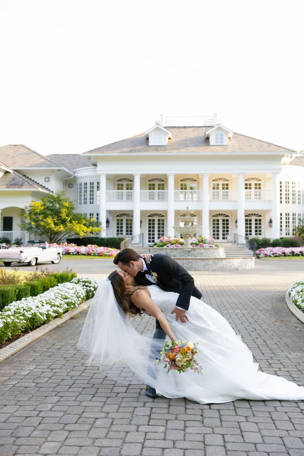 Wedding couple performs dramatic dip pose on stone driveway in front of grand white estate.
