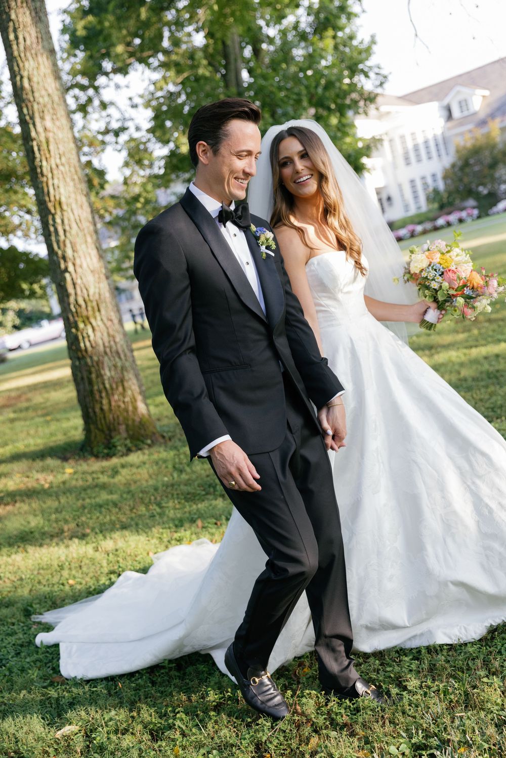 A couple in wedding attire shares a playful moment together on grassy grounds with trees in the background.