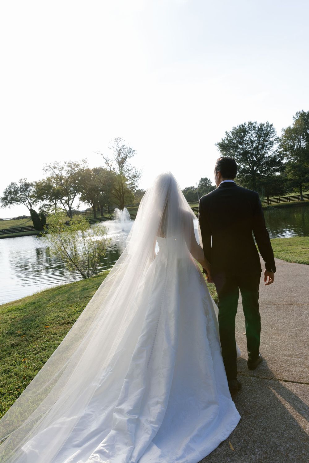 Newlywed couple walking along a scenic lakeside path with flowing white wedding dress trailing behind them.