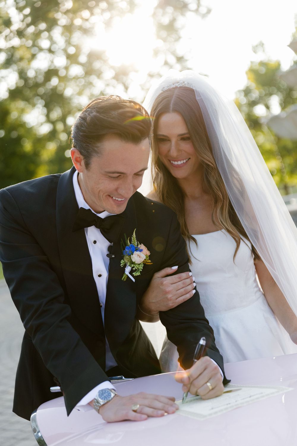 A couple laughs together while signing their marriage certificate outdoors on a sunny day.