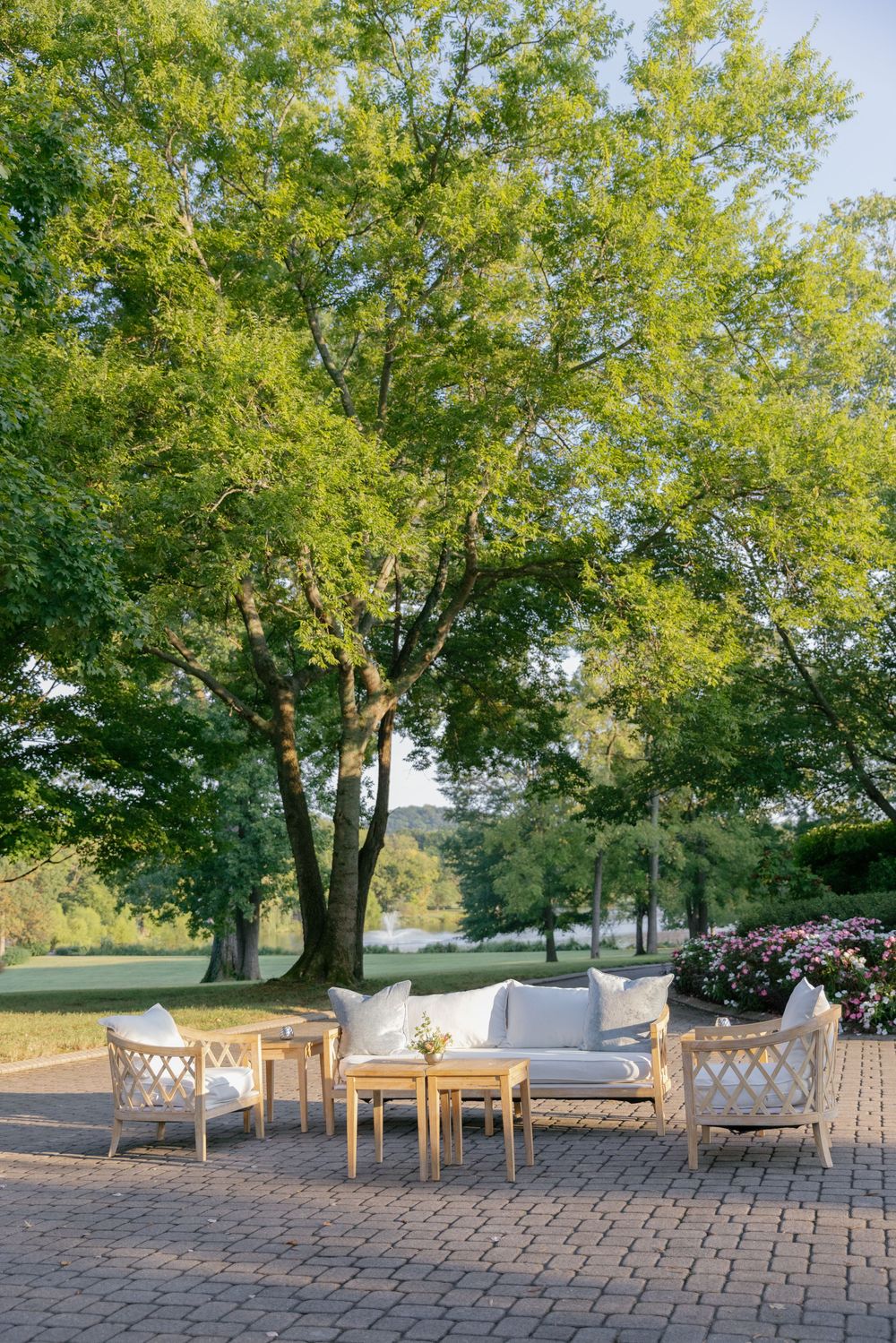 Rustic outdoor wedding venue setup with wooden benches and tables arranged on a stone patio under large green trees.
