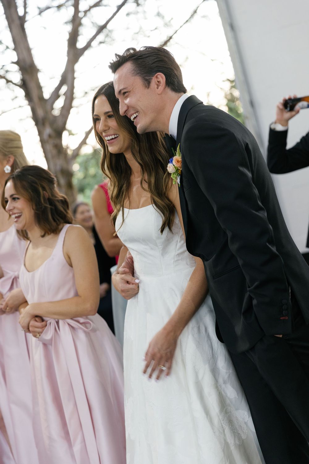 Wedding party in pink bridesmaid dresses and black tuxedos sharing joyful laughter during outdoor celebration.