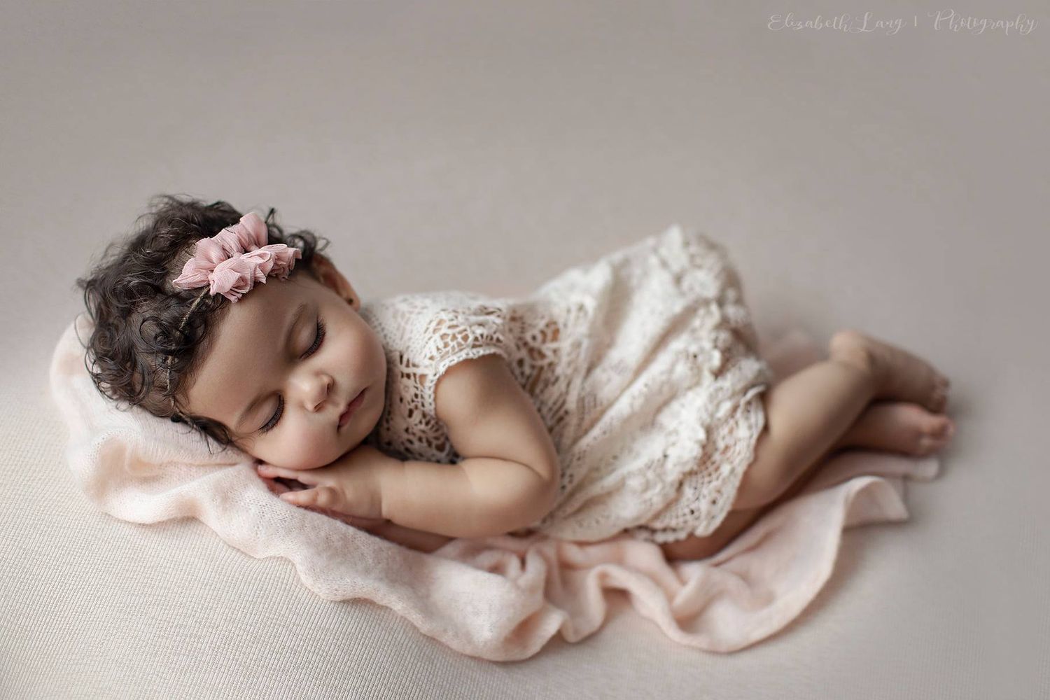 A sleeping newborn baby wrapped in soft pink fabric lies peacefully in a dreamy studio setting.