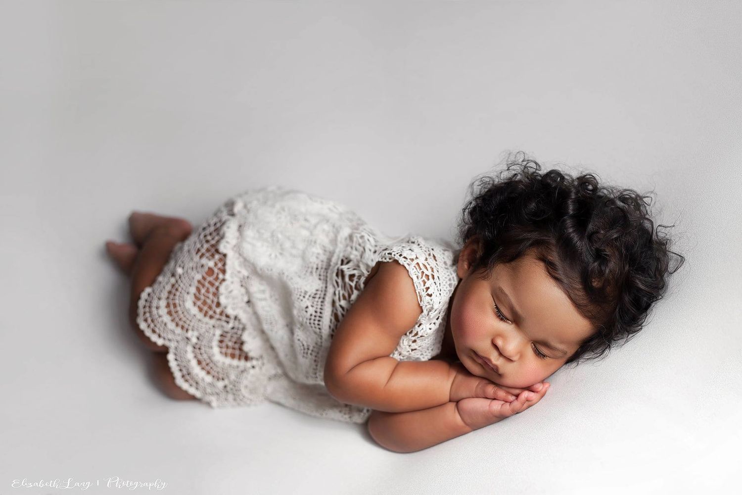 Sleeping baby in white lace dress lies curled up on soft gray backdrop in peaceful newborn photo session.