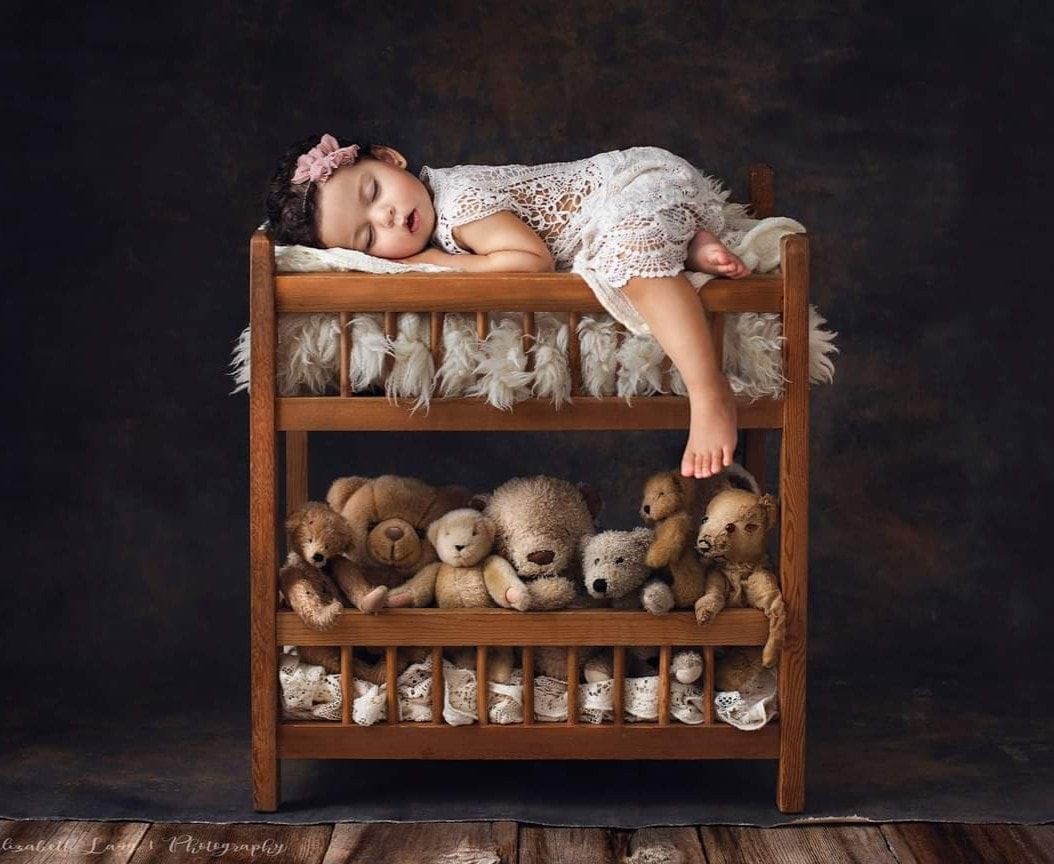 A newborn baby sleeping on a wooden bed with stuffed teddy bears arranged on the shelf below.
