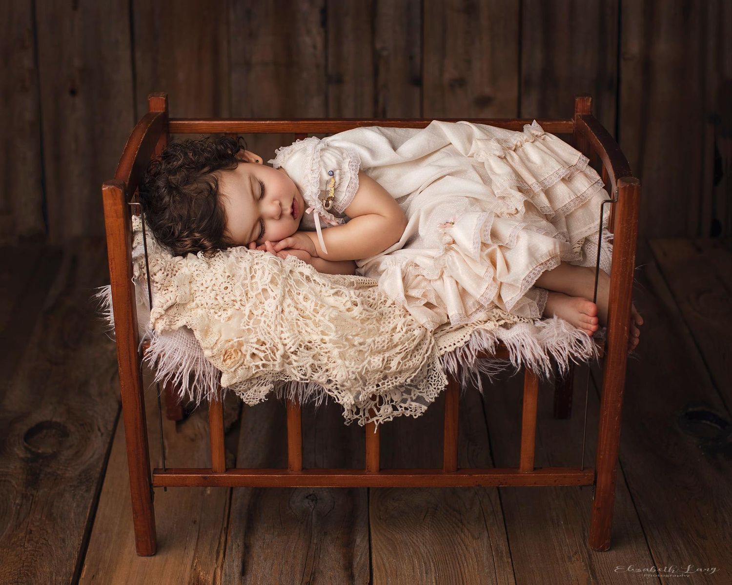 Sleeping baby in vintage lace dress lying on wooden bench in rustic setting.