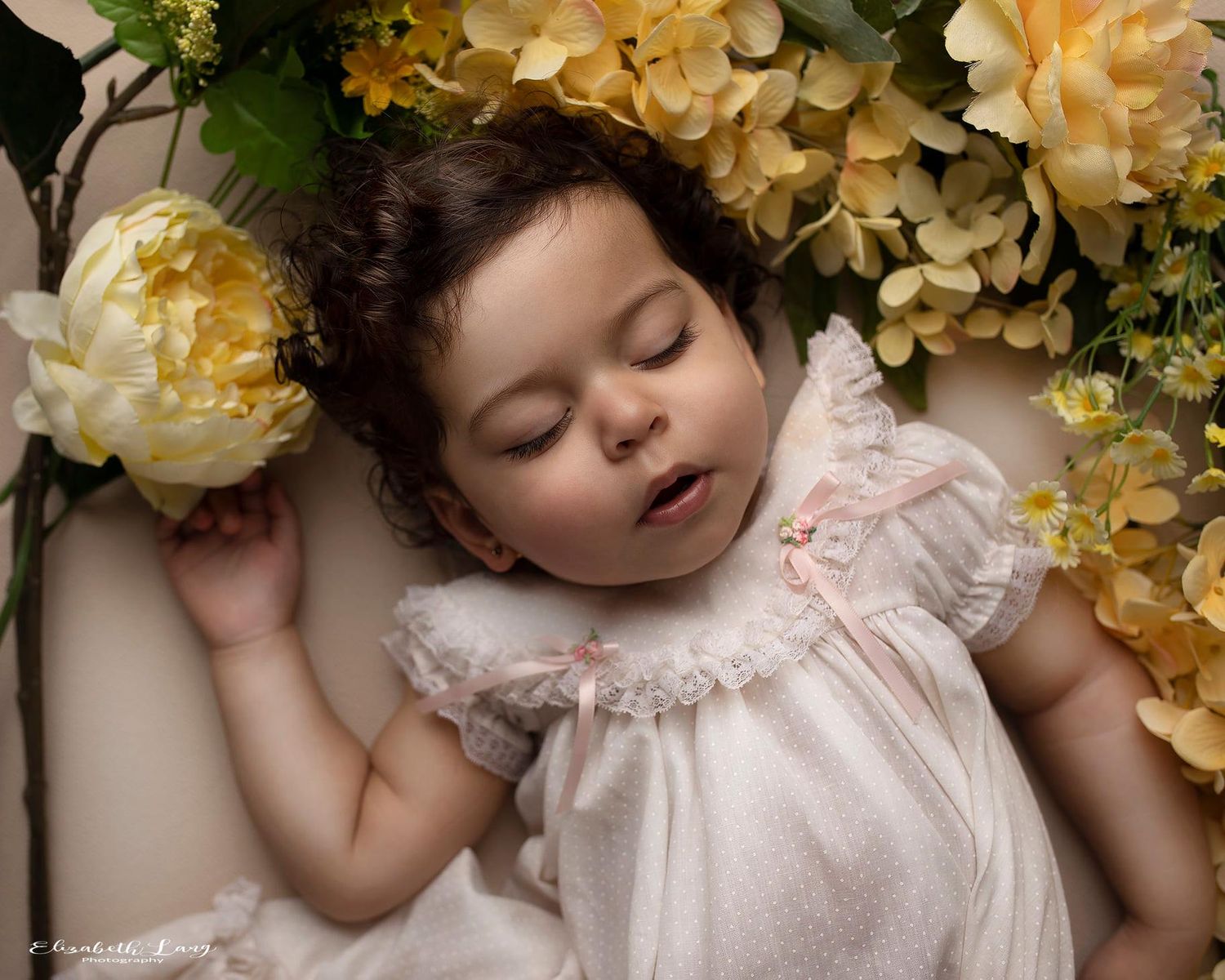 Sleeping baby in pink dress surrounded by cream hydrangeas and floral arrangements.