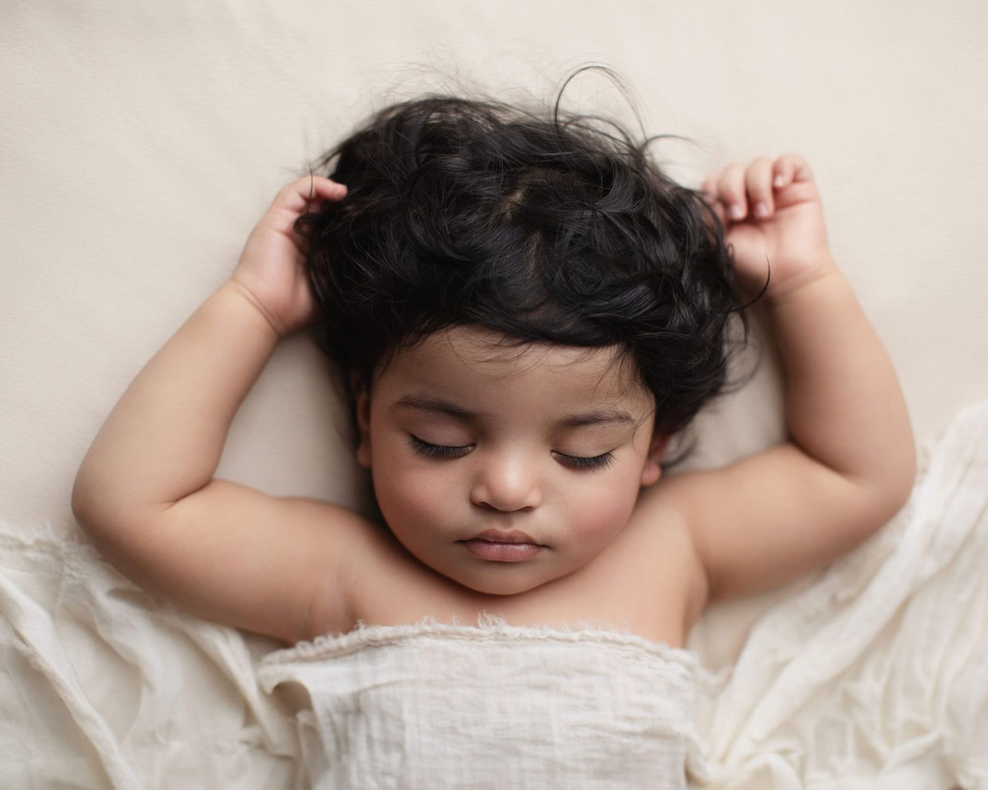 A sleeping baby with dark curly hair rests peacefully on white bedding with arms raised above their head.