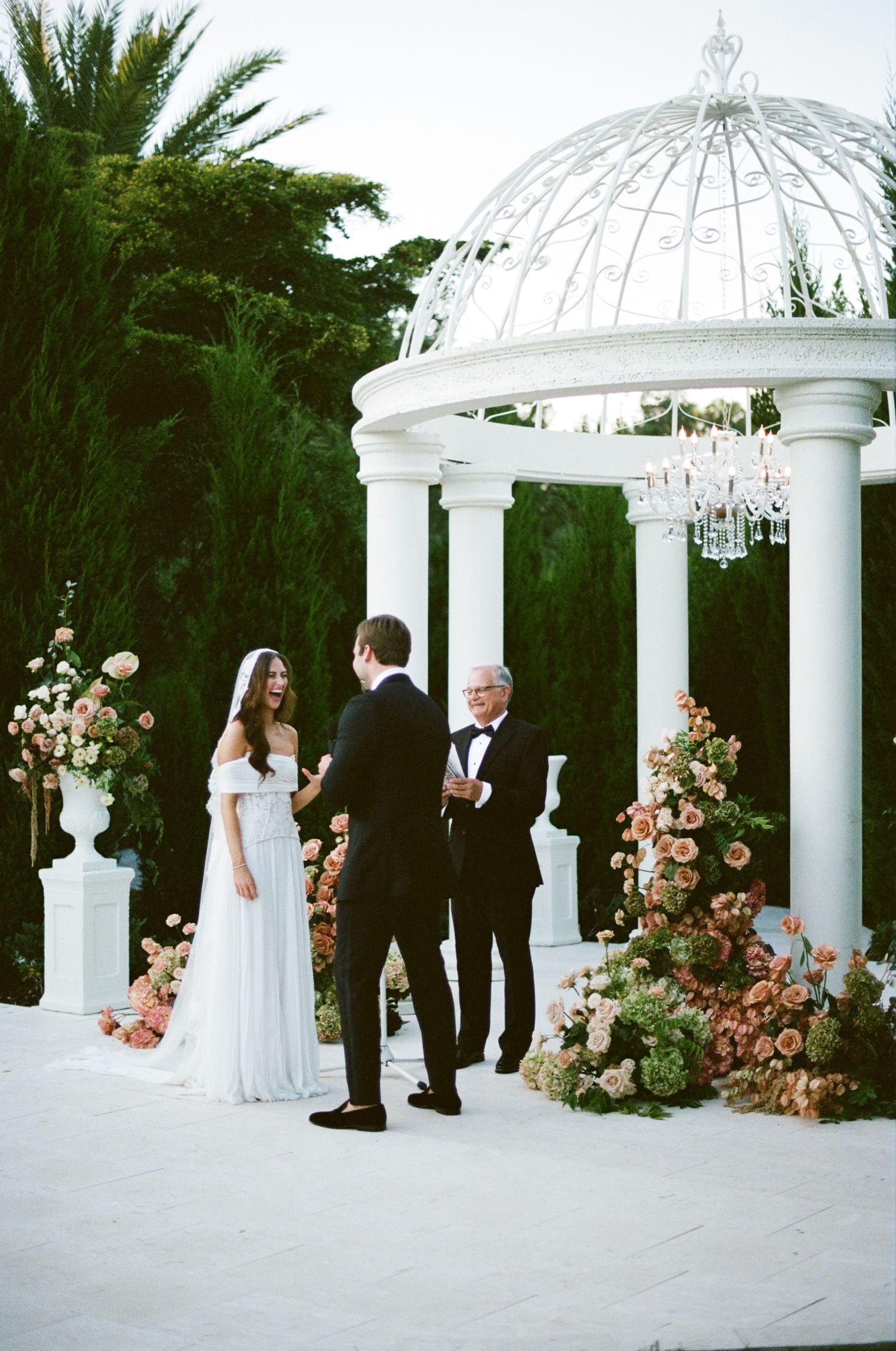 Elegant white gazebo decorated with pink floral arrangements serves as backdrop for outdoor wedding ceremony.