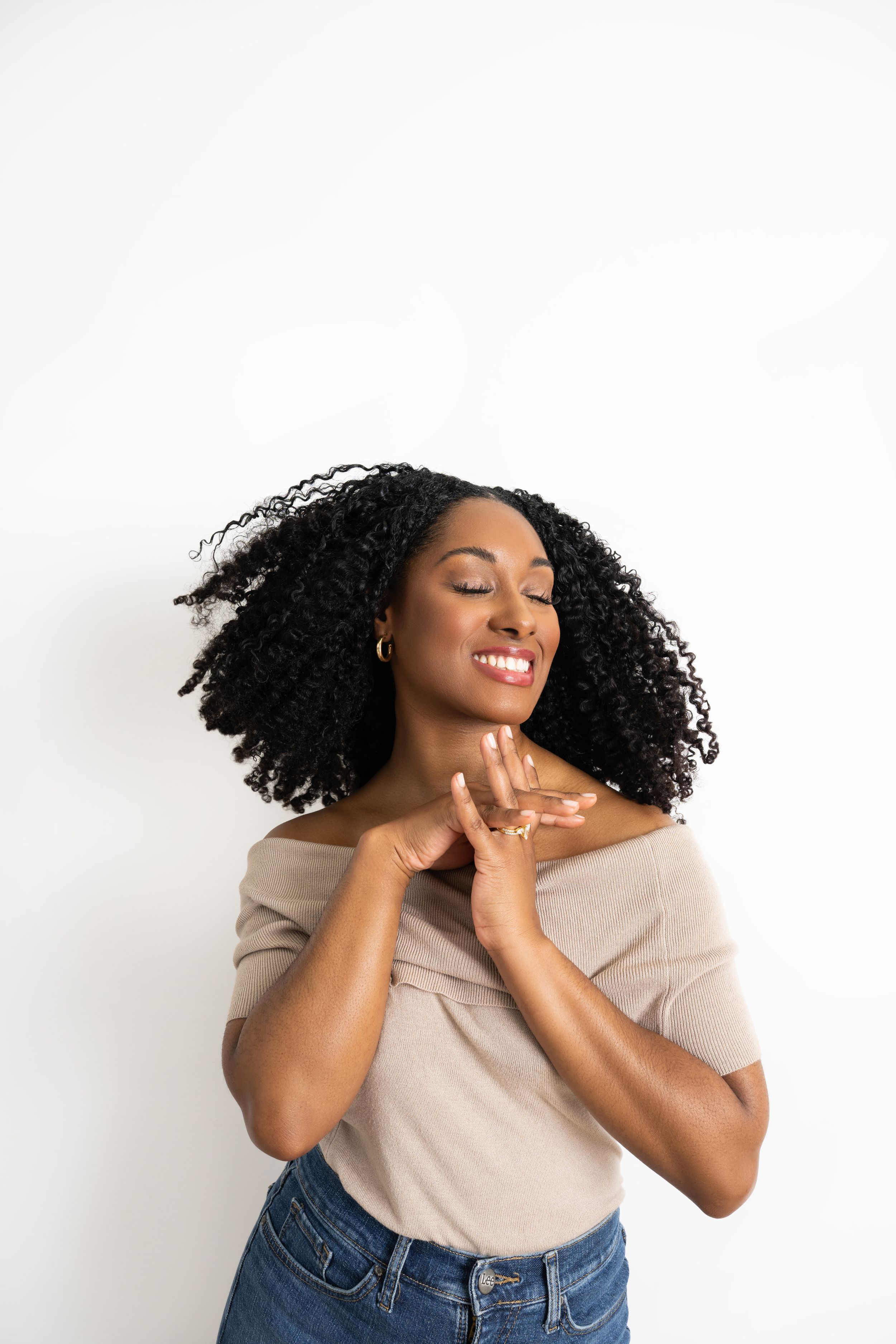 Woman in beige top and jeans poses joyfully against white background with natural curly hair.