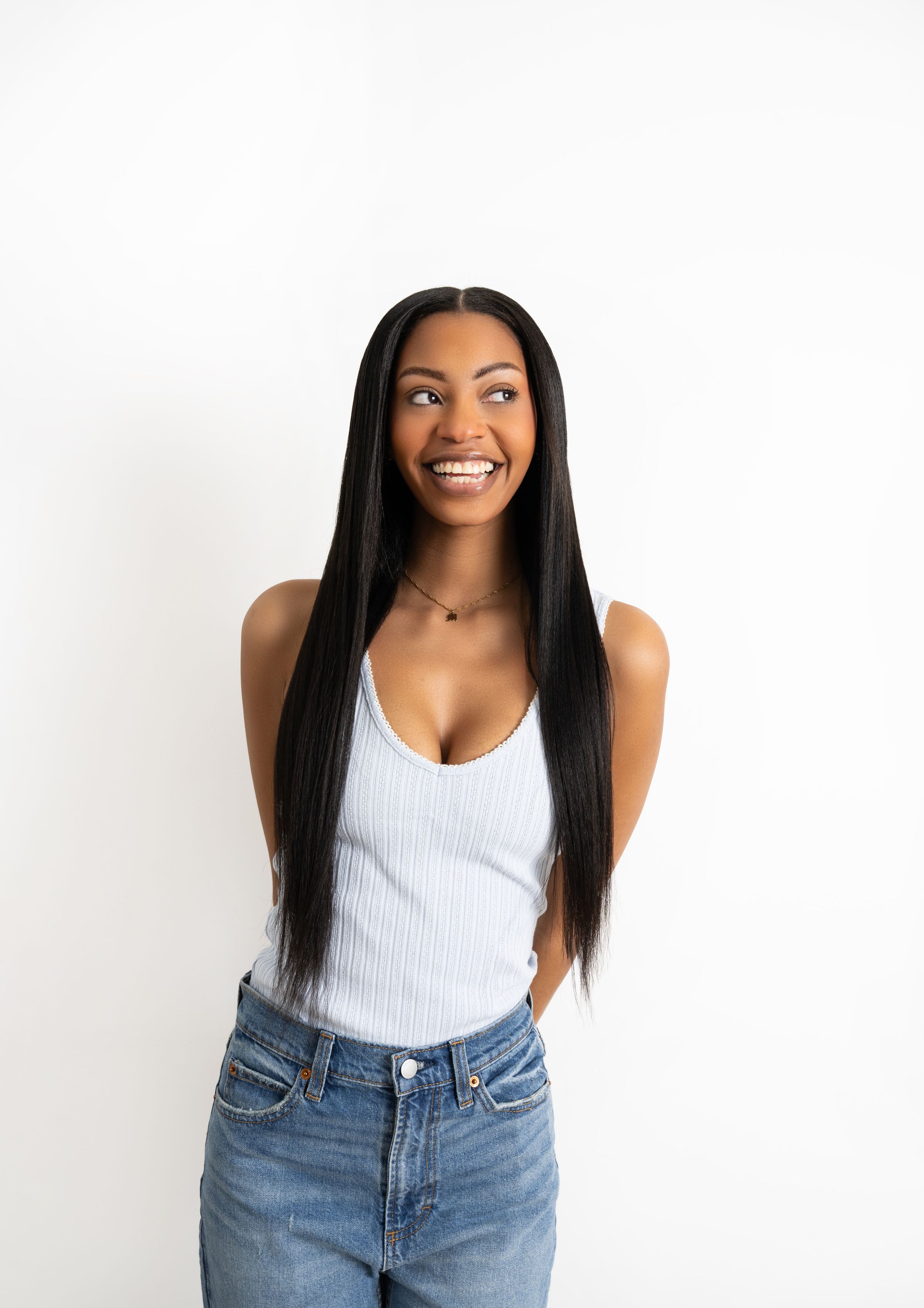 Woman in white tank top and high-waisted jeans smiling against white background.