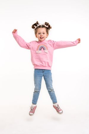 Young child in pink sweatshirt and jeans jumping playfully against white background.