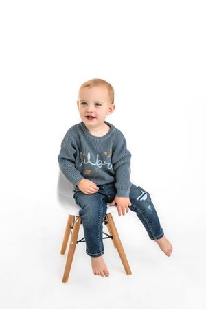Toddler in blue sweater and jeans sitting on a chair in a white backdrop studio for personailty photo session.