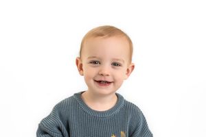 Young Child in blue ribbed sweater smiles while seated against white backdrop for personailty mini sessions.