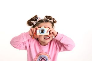 Child in pink sweater pretends to take a photo with a toy camera with hair in buns against a white background.