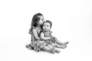 Big sister kisses baby sister on head while seated on a white studio background.