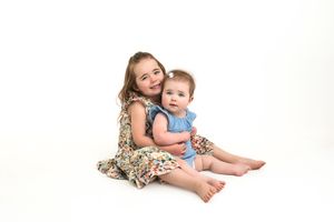 Two sisters cuddling and smiling while sitting on a white studio backdrop for mini sessions.
