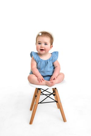 Baby in light blue outfit sits on modern white Eames-style chair against white background.