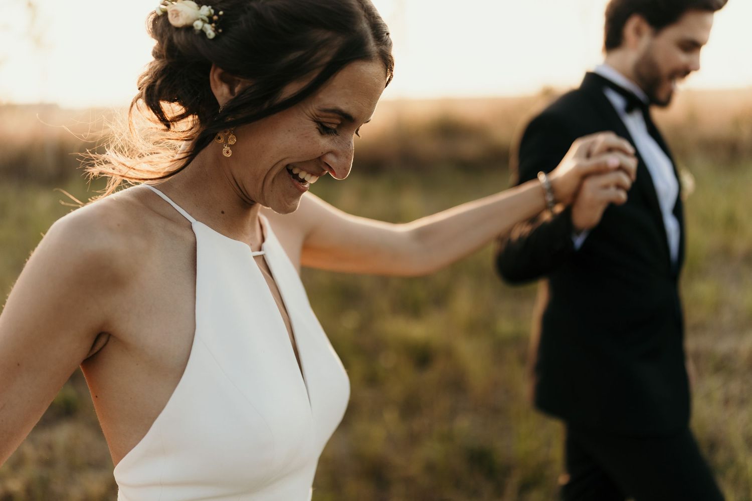 A joyful bride in a white halter dress dances while her groom watches in the background.