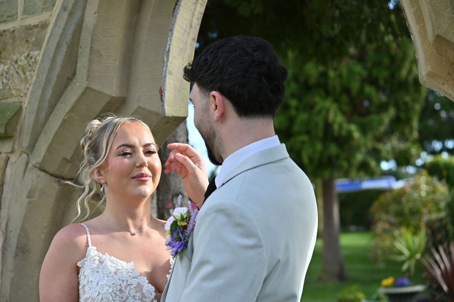 A couple shares a tender moment under an ornate stone archway during their outdoor wedding ceremony.