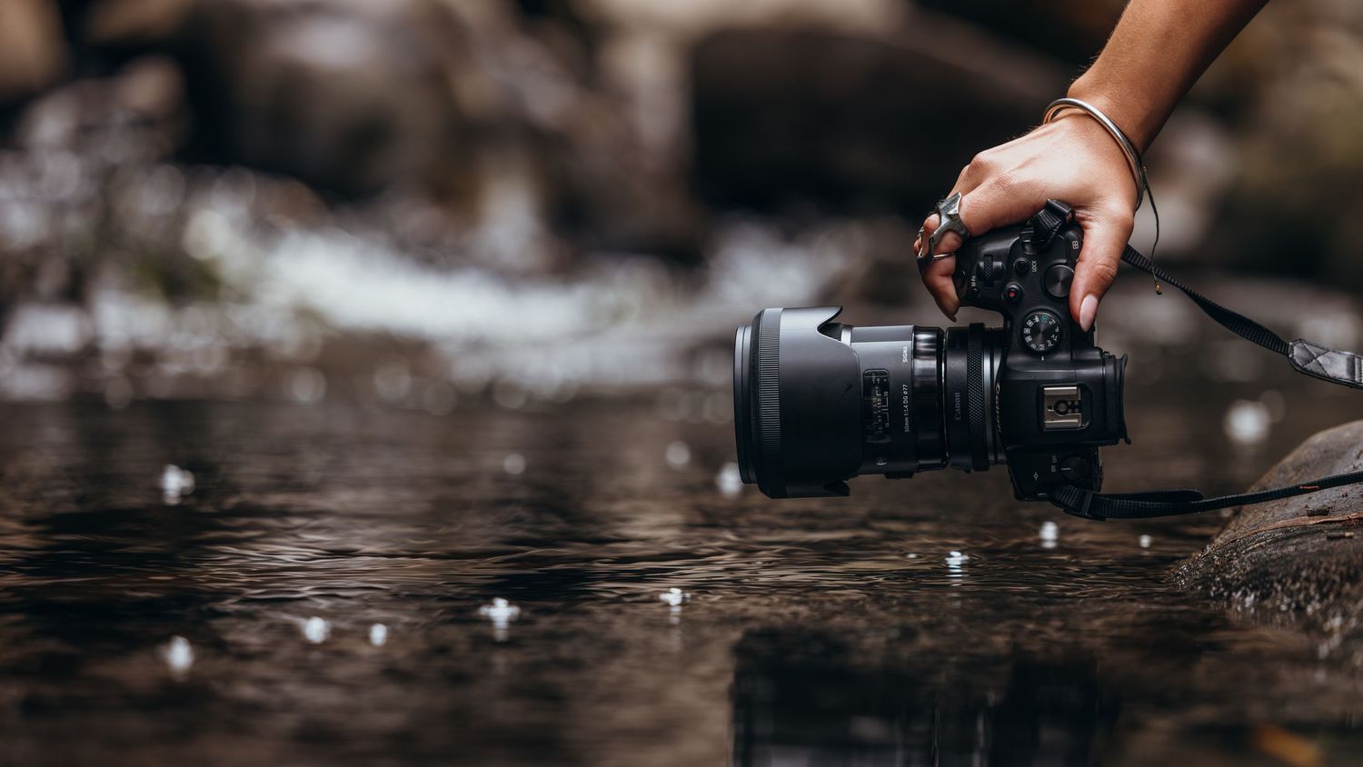 Hand holding professional camera with telephoto lens over reflective water surface.
