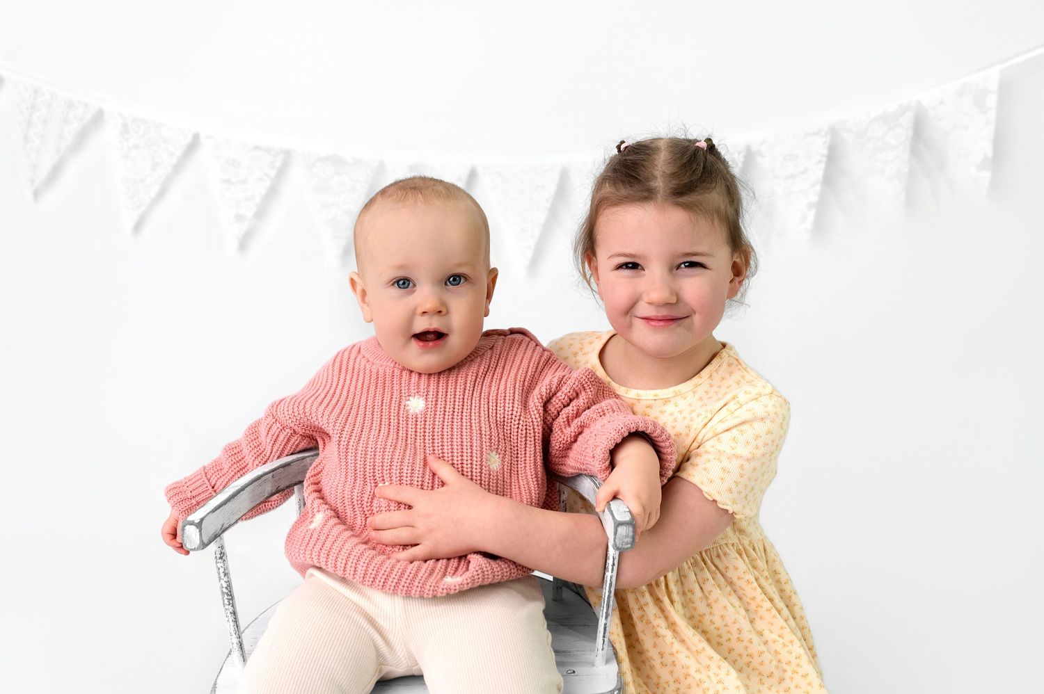 Baby in pink outfit crawls on white backdrop while holding plush teddy bear during studio photo session.