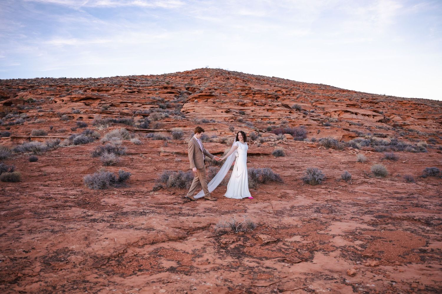 Authentic Des Moines IA wedding couple strolling together in elegant attire against stunning red rock formations at sunset, documentary styl