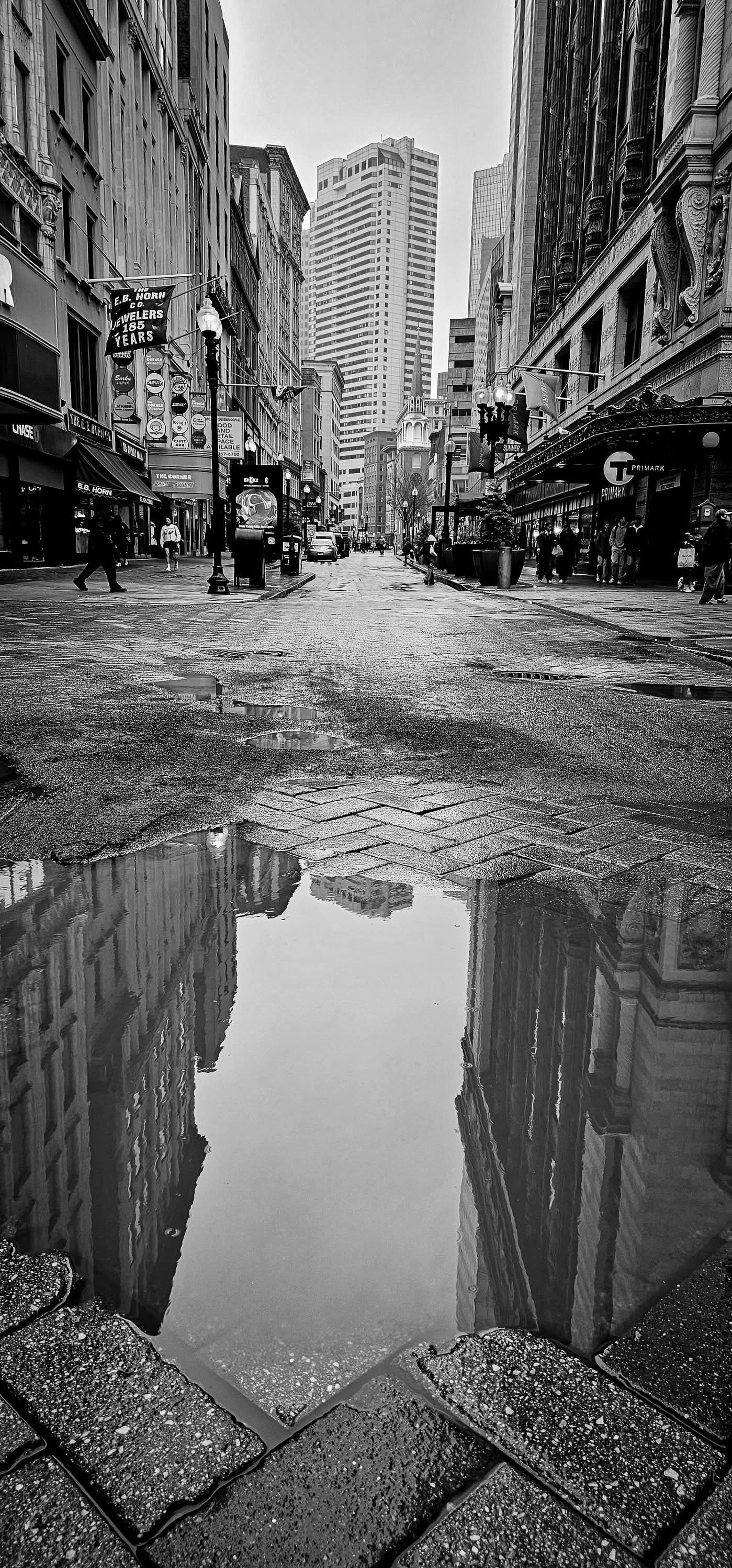 Black and white urban street scene with tall buildings reflected in puddle on wet pavement.