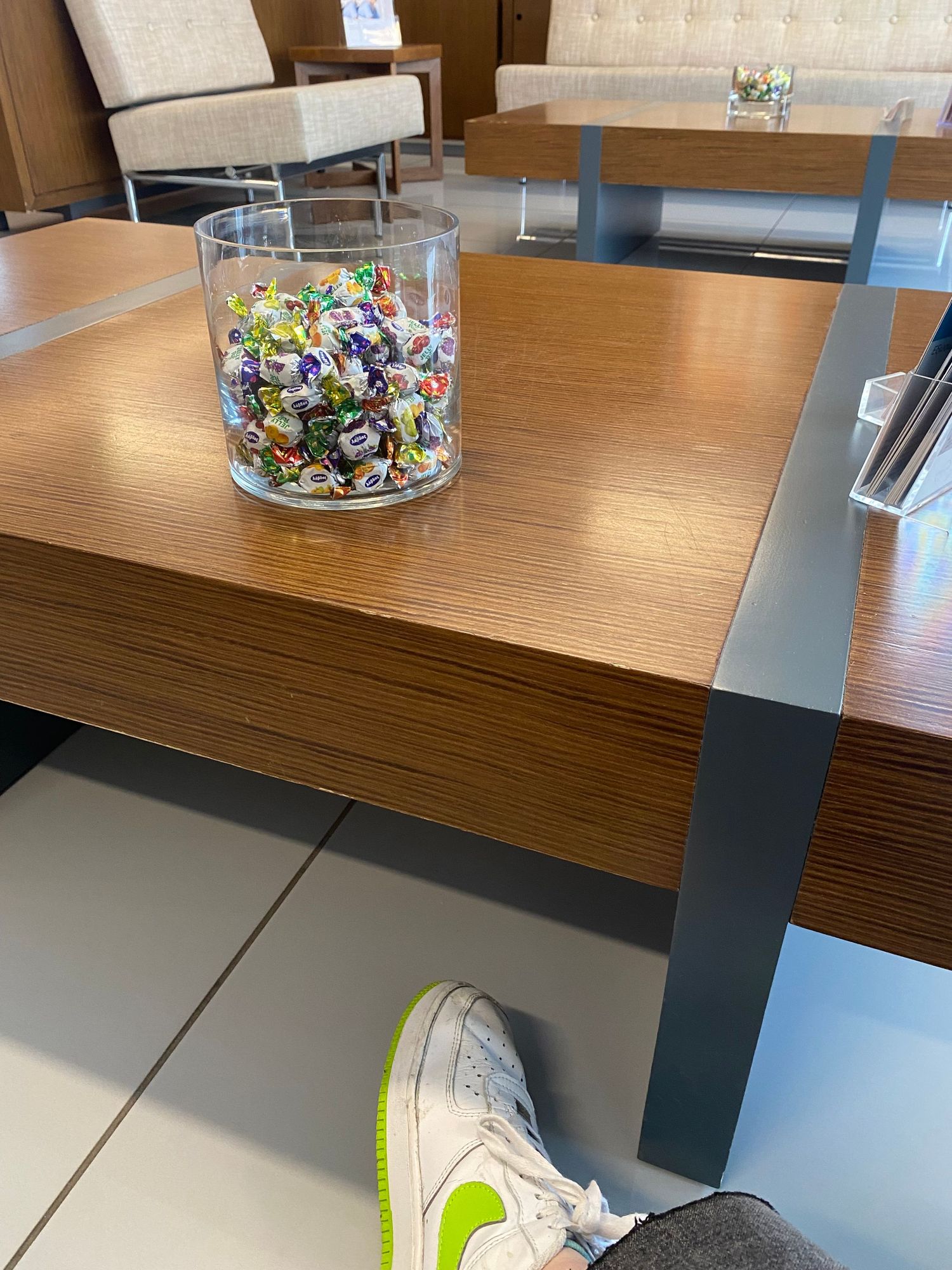 White Nike sneaker with neon green accents sits on a wooden desk next to a glass bowl filled with colorful candies.